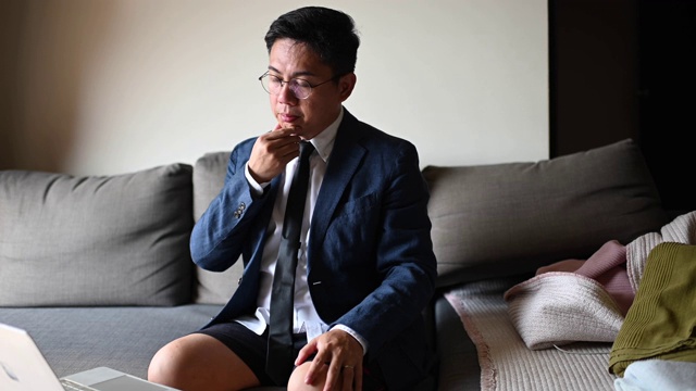 一位打着领带、穿着西装、穿着短裤的亚洲华人中年男子坐在客厅的沙发上，用笔记本电脑与他的商业伙伴和同事进行虚拟商务会议视频下载