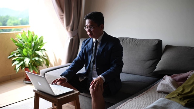 一位打着领带、穿着西装、穿着短裤的亚洲华人中年男子坐在客厅的沙发上，用笔记本电脑与他的商业伙伴和同事进行虚拟商务会议视频下载