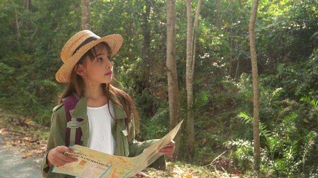 正面:一个十几岁的女孩带着长头发戴着帽子使用和检查地图作为导航，以远足在镜头费日落。儿童背包客在冒险活动的概念。视频素材
