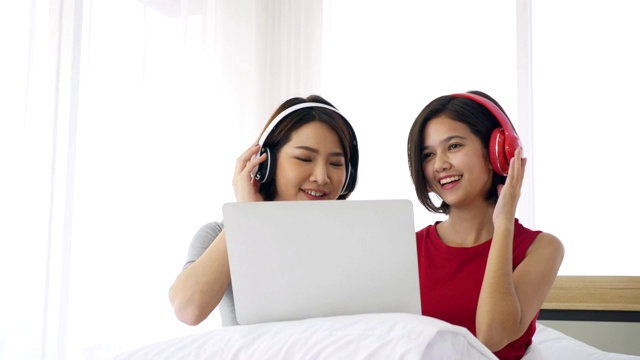 两名亚洲女子戴着耳机在床上用笔记本电脑听音乐。视频下载