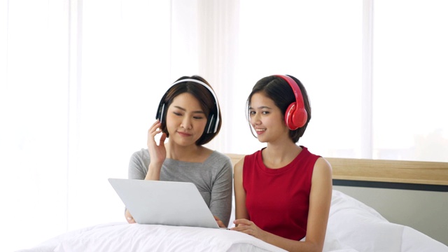 两名亚洲女子戴着耳机在床上用笔记本电脑听音乐。视频下载