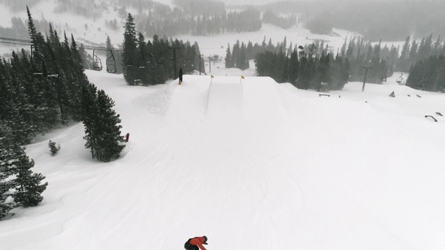 在科罗拉多州博尔德附近的埃尔多拉滑雪度假村，在一个下雪的，阴天完成一个跳跃与森林和滑雪电梯的“前侧360鬼把戏”视频素材