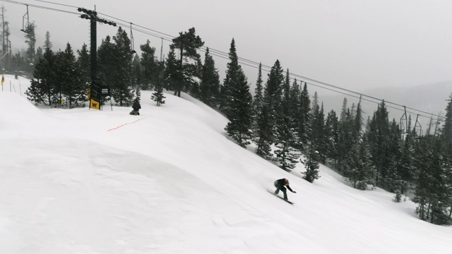 慢动作，无人机拍摄的滑雪板在完整的冬季装备完成一个“背面360法”的伎俩与森林和滑雪升降机在背景在科罗拉多州博尔德附近的埃尔多拉滑雪度假村在一个下雪，阴天视频素材