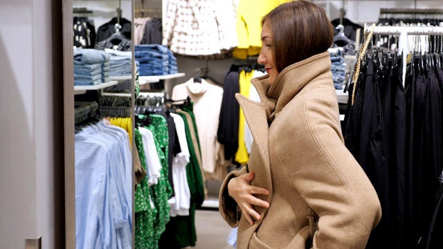 穿着一件温暖的米色羊毛大衣的黑发女人站在一家商店的镜子前视频下载