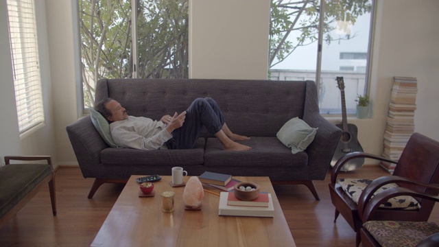 一个成熟的男人躺在沙发上用他的笔记本电脑视频下载