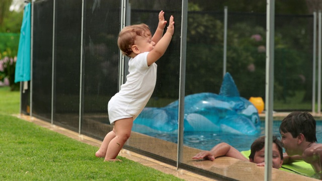 宝宝看着孩子们在夏天的时候在游泳池的水里玩耍。婴儿倚在泳池围栏上视频素材