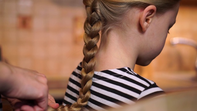 母亲双手精心为女儿编了个辫子和编织的发型，长长的金发女孩穿着条纹裙在厨房舒适的家里。母亲的一天视频下载