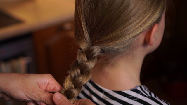 关怀的母亲用手编织的发型编织女儿孩子长发女孩穿着条纹衣服在木制厨房舒适的家。妈妈关心。母亲的一天视频下载