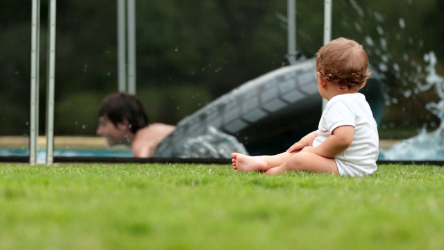 可爱的宝宝坐在后院的草地上，看着孩子们在游泳池里玩耍视频素材