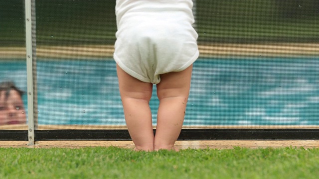 可爱的宝宝站在泳池围栏旁安全，宝宝看着孩子们在水里玩耍视频素材
