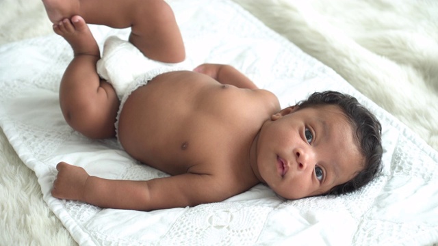高角镜头:微笑的非洲男婴，天真无邪的男婴，带着宠爱躺在卧室的白色床上，带着怀疑的心情看着镜头。父母只有一个孩子的概念。视频素材