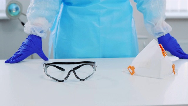 一个眼镜和呼吸器在医院的白色桌子上的特写。背景是一位戴着防护手套和防护服的医生，双手斜靠在桌子上。摄影机慢慢移到一边视频下载