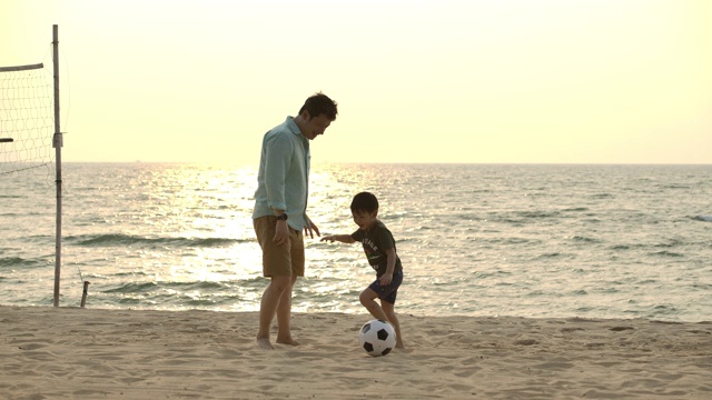 亚洲父亲和儿子一起在沙滩上玩足球在夕阳的背景下。缓慢的运动。幸福的家庭的概念。视频素材