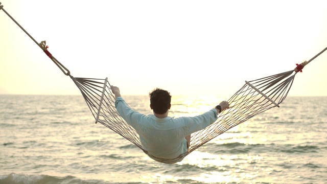 放松的男游客享受他的暑假。年轻的亚洲男子放松在吊床上的海滩和看向地平线享受日落的观点。视频素材