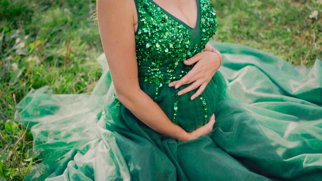 夏天风景如画的一片多汁的草地上，一个年轻的怀孕的女孩正坐在草地上，穿着绿油油的长裙，绿油油的闪闪发光，快乐的时尚模特用双手小心地捧着她的肚子视频素材