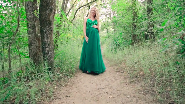 一个年轻的怀孕的已婚金发女人独自走在夏日的森林里，沿着一条狭窄的小路轻轻地走着，她穿着一件漂亮的镶着亮片的祖母绿裙子，用手轻轻地抚摸着她的肚子视频下载