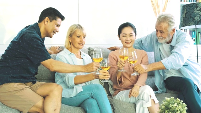 一群老年人在客厅聚会，喝着白葡萄酒，互相敬酒。白人，亚洲人，泰国人坐在家里的沙发上。退休生活方式的概念。视频素材