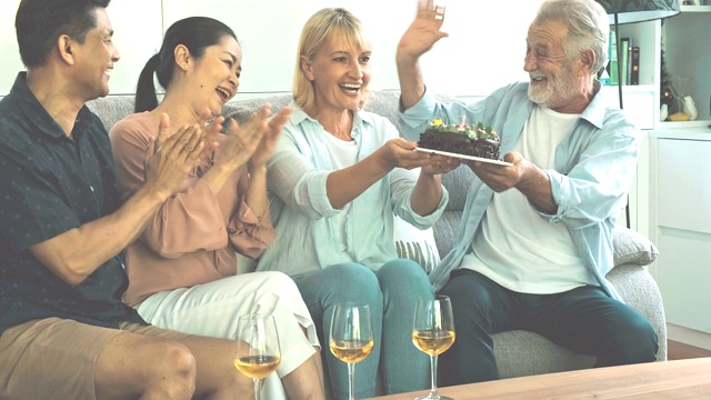 老女人在客厅聚会上吹生日蜡烛。白人，亚洲人，泰国人坐在家里的沙发上。退休生活方式的概念。视频素材