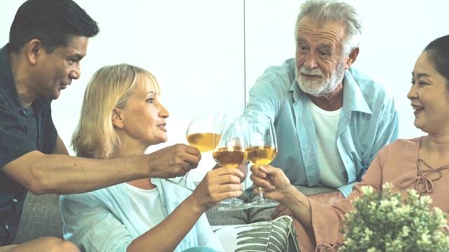 一群老年人在客厅聚会，喝着白葡萄酒，一起庆祝。白人，亚洲人，泰国人坐在家里的沙发上。退休生活方式的概念。视频素材