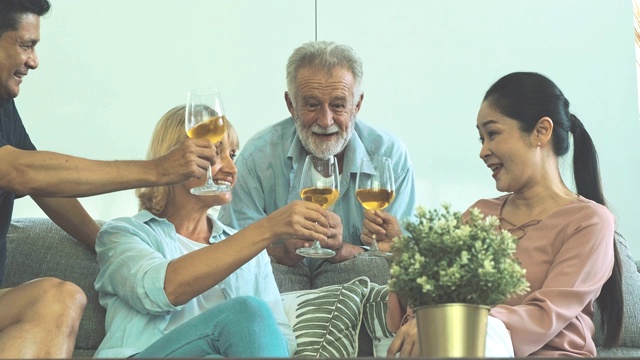 一群老年人在客厅聚会，喝着白葡萄酒，一起敬酒。白人，亚洲人，泰国人坐在家里的沙发上。退休生活方式的概念。视频素材