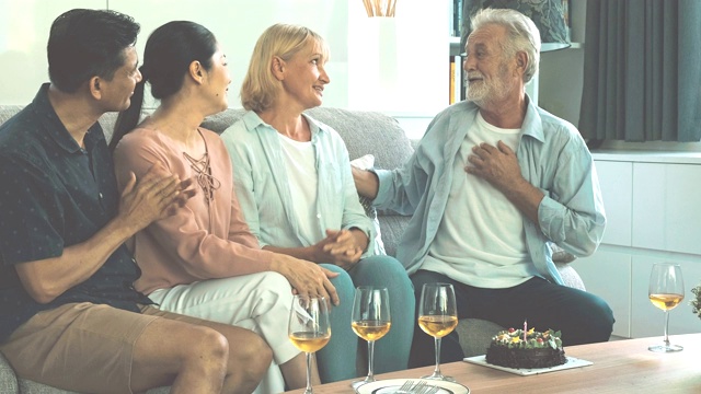一对老夫妇在生日聚会上和朋友在客厅拥抱。白人，亚洲人，泰国人坐在家里的沙发上。退休生活方式的概念。视频素材