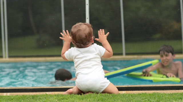 刚学会走路的婴儿站在游泳池的安全围栏旁，孩子们在夏天玩耍视频素材