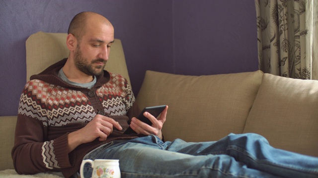 一个年轻人坐在家里的沙发上看电子书视频素材