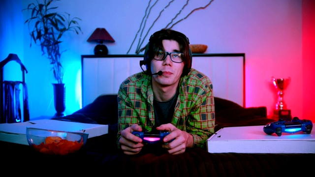 年轻英俊的男子戴着耳机躺在床上玩在线视频游戏，对着麦克风说话视频下载
