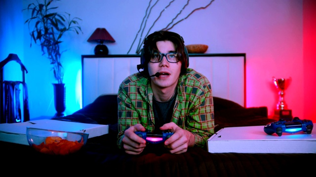 年轻英俊的男子戴着眼镜和耳机躺在床上玩在线视频游戏视频下载