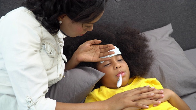 4K非洲裔美国母亲用手和数字口腔温度计检查生病的女儿的温度，并把发烧贴在她的头上。可爱的小女孩躺在床上患了感冒和流感。母亲和保健概念。视频素材