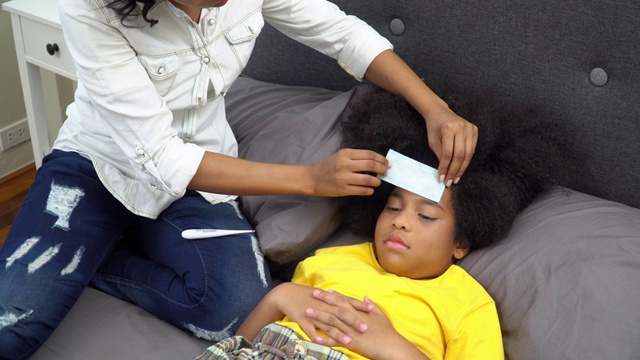 4K非洲裔美国母亲把发烧贴在生病的女儿头上，并使用数字口腔温度计检查温度。可爱的小女孩躺在床上患了感冒和流感。母亲和保健概念。视频素材