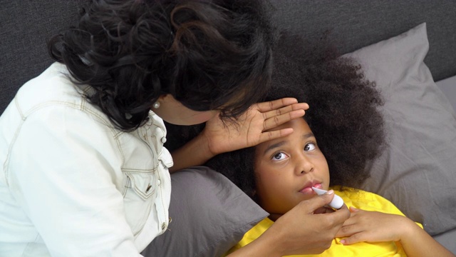 4K非洲裔美国母亲用手和数字口腔温度计检查她生病的女儿的温度。可爱的小女孩躺在床上患了感冒和流感。母亲和保健概念。视频素材