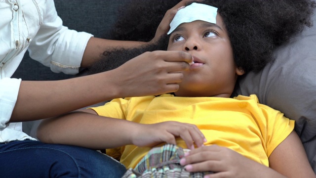 4K非洲裔美国母亲把发烧贴在生病的女儿头上，并使用数字口腔温度计检查温度。可爱的小女孩躺在床上患了感冒和流感。母亲和保健概念。视频素材