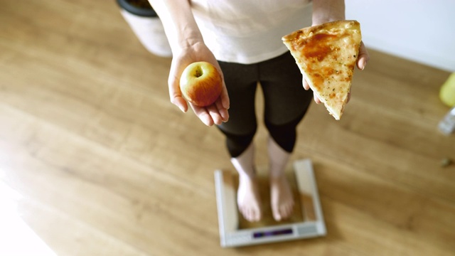 SLO MO拍摄了一个面目人非的女人一手拿着一片披萨，一手拿着一个苹果踩在体重秤上的照片。视频下载
