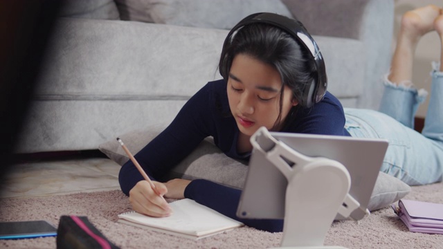 迷人的亚洲少女戴着耳机听和学习在平板电脑屏幕上与她的老师，并在笔记本上写冠状病毒或冠状病毒19封锁情况。视频电话技术与她的建议学习她的家庭作业。视频素材