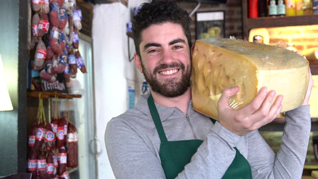 一个快乐的年轻人在一家熟食店工作，他拿着一大块很重的轮式奶酪，对着镜头微笑视频下载
