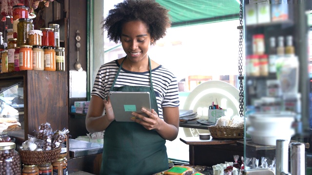 美丽的黑人妇女在一家熟食店工作，看着一边沉思一边拿着一个平板电脑视频下载