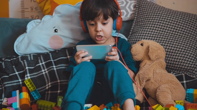 5岁男孩在玩手机游戏。学龄前儿童在小工具上玩游戏。他使用耳机以避免受到干扰。视频素材