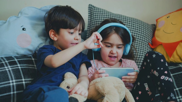 兄弟姐妹一起玩手机游戏。男孩教他的妹妹玩。快乐地玩游戏。使用耳机以避免受到干扰。一对五岁的双胞胎在凌乱的沙发上玩耍。学龄前儿童在电子设备上玩游戏。视频素材