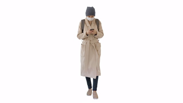 一名妇女穿着一件大衣，戴着医用口罩，用手机和行走在白色的背景视频下载