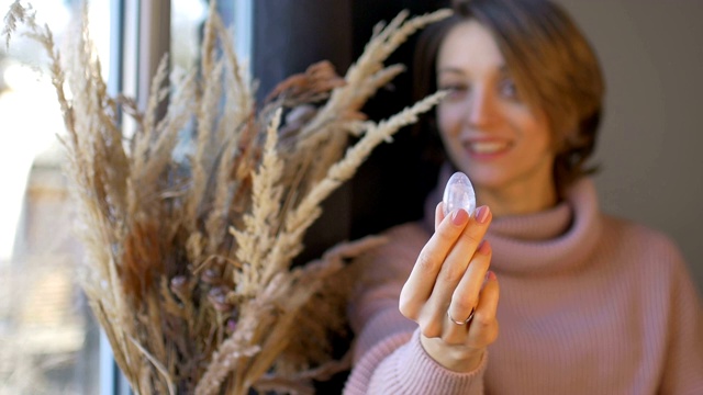 一个女人拿着由透明的紫水晶制成的女性卵子，站在窗前，旁边放着一花瓶的小穗。女性健康理念、养生、修身或冥想视频下载