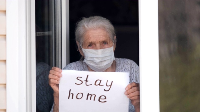 一位戴着医用口罩的灰白头发的老妇人站在家里靠窗的地方，手里举着一张写着“待在家里”的海报。视频下载