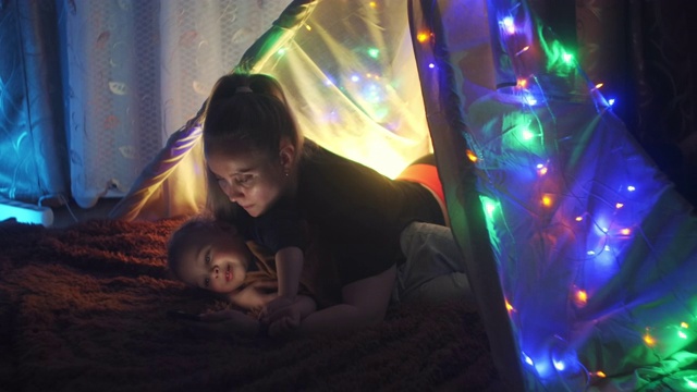 睡前故事。快乐的妈妈和小儿子在家里的帐篷里看书。晚上在黑暗的房间里，用手电筒盖着毯子看书。白人妇女享受与儿子的家庭时光。视频素材