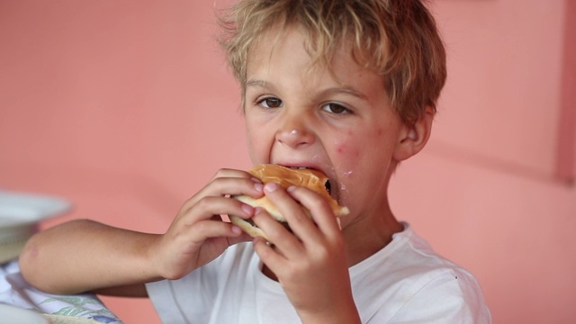 可爱的小男孩在吃汉堡包。金发男孩吃了一口汉堡视频下载
