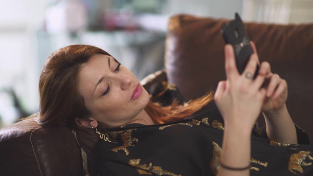在法国covid-19大流行期间，迷人的红发女子在家里的棕色沙发上发短信。一个年轻女孩使用智能手机。视频下载