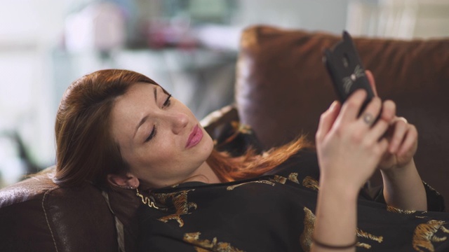 在法国covid-19大流行期间，迷人的红发女人微笑着，坐在家里的棕色沙发上。一个年轻的女孩正在用智能手机打电话。视频下载