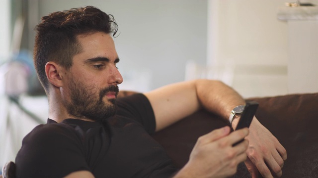 在法国covid-19大流行期间，一名白人男子在社交媒体上使用智能手机，身穿黑色衬衫，坐在家里的棕色沙发上。一人青年孤立无援。视频下载