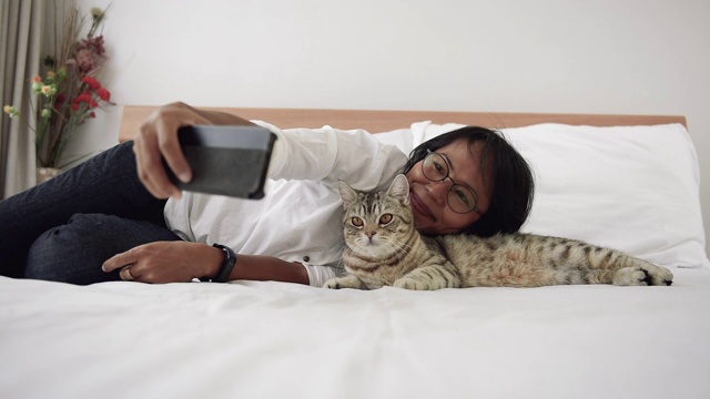 亚洲女性使用智能手机自拍和玩虎斑猫在现代卧室。视频下载