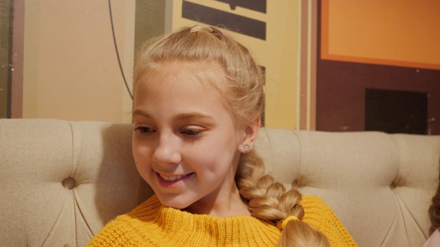 可爱的微笑少女与金发辫子使用移动在沙发上。肖像美丽的女孩看智能手机屏幕上的家庭沙发。视频下载