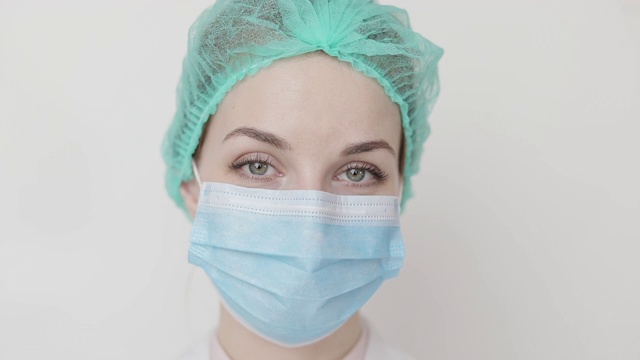 一个女性外科医生的脸的特写，在医疗帽和面具，在镜头眨眼。医务专业女医生护士戴着磨砂帽，防护面膜，对着镜头眨眼视频素材